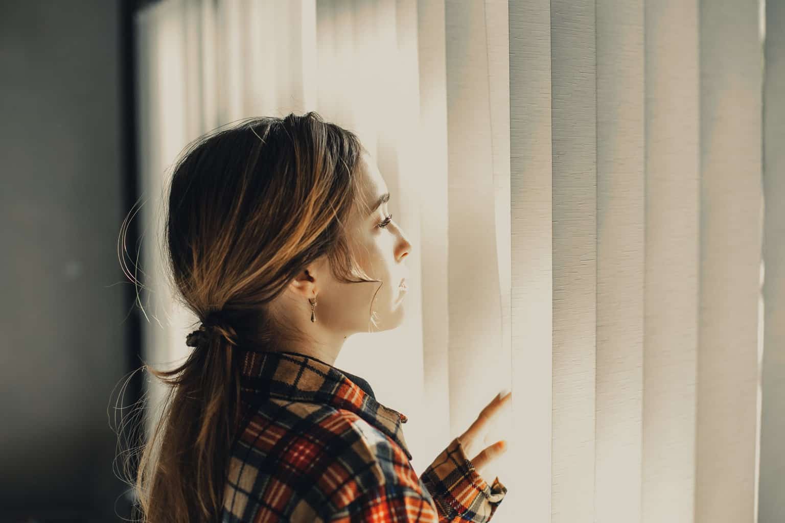 thoughtful young woman looking through blinds of window in daylight