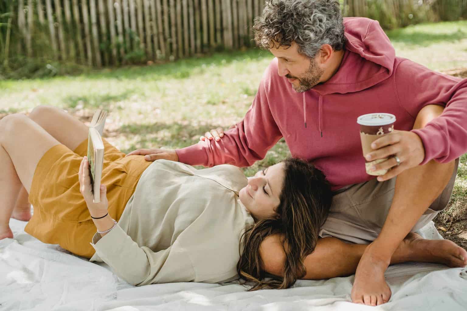 happy pregnant couple reading book and drinking coffee during picnic on lawn